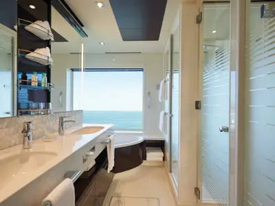 Ein elegantes Badezimmer an Bord der Norwegian Bliss mit zwei Waschbecken, einem separaten Badewannenbereich und einer Duschkabine. Ein großes Fenster bietet einen weiten Blick auf das offene Meer. Handtücher und Pflegeprodukte sind bereitgelegt.