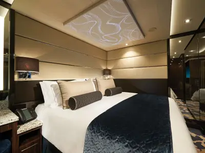 Ein stilvolles Schlafzimmer in der Haven Suite an Bord der Norwegian Bliss, mit einem großen Bett, eleganten Kissen und einer markanten Deckenbeleuchtung. Rechts reflektiert ein Spiegelschrank das Zimmer.