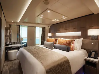 Ein elegantes Schlafzimmer in einer Suite an Bord der Norwegian Bliss mit großem Bett, Nachttischen und einem Schreibtisch. Durch die Balkontür ist das weite Meer sichtbar. Eine gemütliche und moderne Atmosphäre.