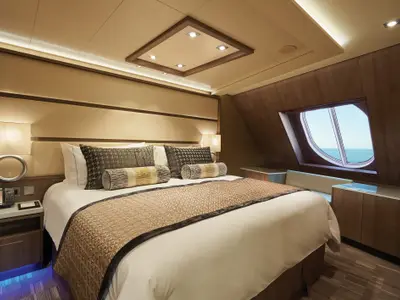 Elegantes Schlafzimmer in einer Suite der Norwegian Bliss. Grosses Bett, Nachttisch mit Telefon. Ein ovales Fenster bietet Meerblick. Warme Beleuchtung.