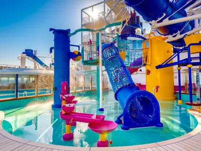 Ein lebhafter Kids Aqua Park auf dem Deck der Norwegian Bliss. Blaue, gelbe und pinke Rutschen sowie Wasserspielgeraete laden zum Spass ein. Wasser spritzt in die hellblaue Poollandschaft unter klarem Himmel.