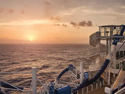Atemberaubender Sonnenuntergang über dem Meer, mit der großen Wasserrutsche an Bord der Norwegian Bliss im Vordergrund. Der Himmel leuchtet in warmen Orangetönen.