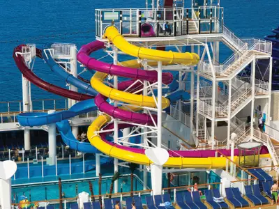 Eine dynamische Ansicht der mehrstufigen Wasserrutschenanlage an Bord der Norwegian Breakaway. Mehrere bunte Röhrenrutschen in Gelb, Magenta, Rot und Blau schlängeln sich um eine weiße Stahlkonstruktion über dem Deck. Im Hintergrund ist das tiefblaue Meer unter hellem Tageslicht zu sehen. Einige kleine Figuren von Personen sind auf den Treppen und am Poolbereich erkennbar.