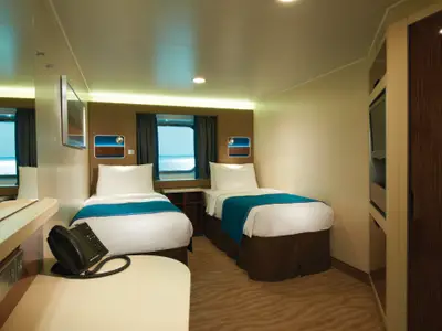 Eine helle und moderne Außenkabine an Bord der Norwegian Breakaway mit zwei Einzelbetten, einem Fenster zum Meer und funktionaler Einrichtung. Eine Telefonanlage steht auf dem Schreibtisch. Der Raum ist warm beleuchtet.
