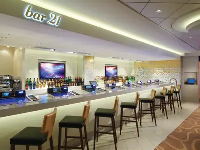 Die Bar 21 an Bord der Norwegian Breakaway zeigt eine lange Theke mit integrierten Bildschirmen und vielen Barhockern, eingerahmt von Spirituosenregalen und leuchtenden TVs.