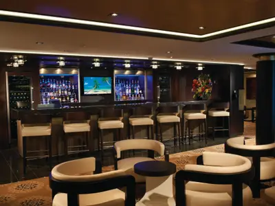 Die stilvolle Bar-Lounge an Bord der Norwegian Breakaway mit langer Theke, Barhockern und beleuchteten Spirituosen. Ein Weinkühlschrank ist sichtbar, ebenso ein Bildschirm mit tropischer Szene. Gemütliche Sessel mit kleinen Tischen laden zum Entspannen ein.