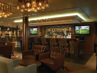 Die Maltings Bar & Whiskey Bar an Bord der Norwegian Breakaway: Eine elegante Lounge mit Ledersesseln, einer gut bestückten Bar und Flachbildfernsehern, die Golfszenen zeigen.