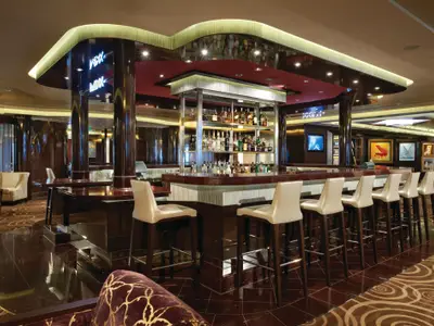 Die elegante Mixx Bar an Bord der Norwegian Breakaway. Im Vordergrund eine lange, glänzende Theke mit hellen Barhockern. Dahinter ein beleuchtetes Spirituosenregal. Dunkle Holzoberflächen und moderne Kunstwerke prägen das luxuriöse Ambiente.