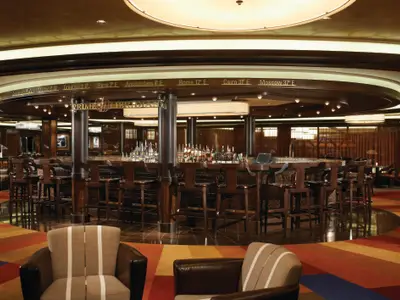 Prime Meridian Bar auf der Norwegian Breakaway. Runder Tresen mit Barhockern, Spirituosen. Weltzeitanzeige mit Städten wie Greenwich 0° über der Bar. Warme Beleuchtung, bunter Teppich.