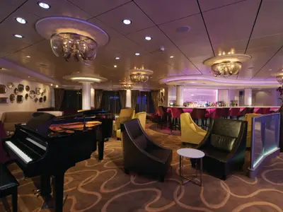 Eine modern und elegant gestaltete Bar-Lounge mit lila Beleuchtung an Bord der Norwegian Breakaway. Ein schwarzer Flügel, bequeme Sessel und eine voll ausgestattete Bar sind zu sehen, sowie eine Wanddekoration aus runden Metallobjekten.