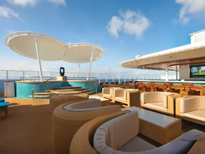 Loungebereich auf dem Deck der Norwegian Breakaway mit Rattan-Sitzgruppen, einer Bar und einem Pool. Im Hintergrund ist das blaue Meer unter klarem Himmel zu sehen. Ein Bildschirm an der Bar zeigt Golf.