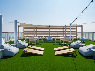 Outdoor leisure deck on Norwegian Breakaway with artificial green turf, cornhole boards, and grey bean bag chairs. String lights and ocean in background. No people.