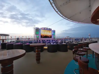 Abendaufnahme des Aussenbereichs Spice H2O an Bord der Norwegian Breakaway. Ein grosser Bildschirm zeigt ein Konzert, umgeben von Loungemöbeln und einer Bar. Das Meer ist im Hintergrund zu sehen.