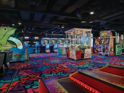 Eine bunte, lebhafte Spielhalle an Bord der Norwegian Breakaway mit Big Bass Wheel Pro, Guitar Hero Arcade und Greifautomaten. Der auffällige Teppich hat ein mehrfarbiges Muster.