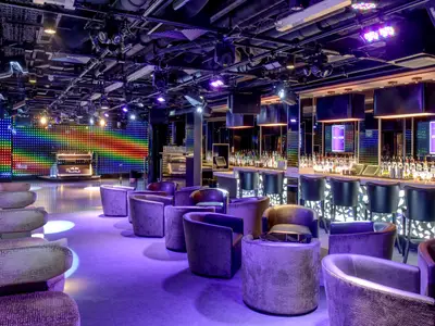 Der Club Bliss an Bord der Norwegian Breakaway: Eine Bar mit Flaschen, Hockern, viele Sessel und eine DJ-Bühne mit bunter LED-Wand. Moderne, lebhafte Atmosphäre mit violetter Beleuchtung.