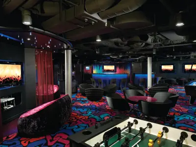 Der lebhafte Club Entourage an Bord der Norwegian Breakaway. Ein dunkler Raum mit farbenfrohem Teppich, blauer Bar, bequemen Sitzecken, Flachbildschirmen, Billard- und Kickertisch.