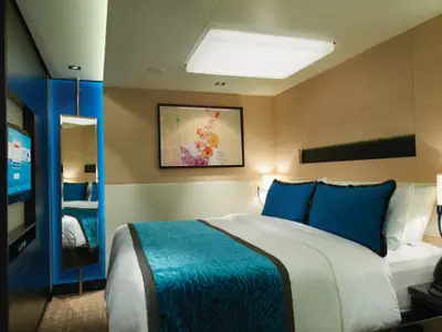 Gemütliche Innenkabine an Bord der Norwegian Breakaway mit Doppelbett, blauen Kissen und teal-farbenem Läufer. Moderne Einrichtung mit TV, Spiegel und Kunst an der Wand.