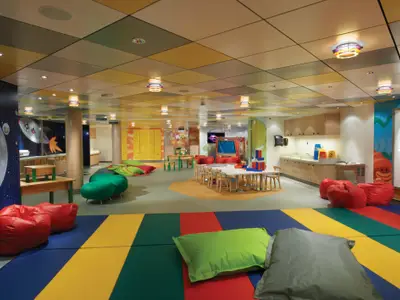 Der farbenfrohe Kinderbereich Splash Academy an Bord der Norwegian Breakaway. Er zeigt eine weite Fläche mit Spielmatten, roten Sitzsäcken und Bereichen mit Tischen für kreative Aktivitäten. An den Wänden sind thematische Illustrationen und ein Spülbeckenbereich zu sehen, während die Decke mit bunten Kacheln und besonderen Leuchten gestaltet ist.