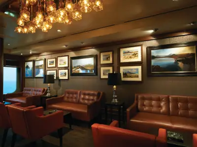 Eine elegante Lounge an Bord der Norwegian Breakaway. Sie zeigt braune Ledersofas, kleine Tische und gerahmte Landschaftsbilder. Ein Fenster bietet Meerblick.