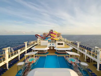 Von oben aufgenommenes Deck der Norwegian Breakaway mit AquaPark, Hauptpool und Whirlpools. Bunte Wasserrutschen, umgeben von vielen leeren blauen Liegestühlen, blicken aufs offene Meer.