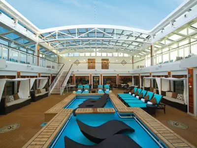 Eine Weitwinkelaufnahme des stilvollen, überdachten Poolbereichs The Haven an Bord der Norwegian Breakaway, mit zwei Pools, bequemen Liegen, privaten Cabanas und umliegenden Restaurants. Tageslicht fällt durch das Glasdach.