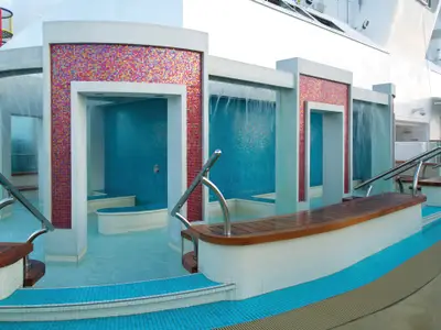 Der moderne Spice H2O Bereich an Bord der Norwegian Breakaway mit Wasserfällen, türkisfarbenen Pools, Holzbänken und farbenfrohen Mosaikwänden unter freiem Himmel.