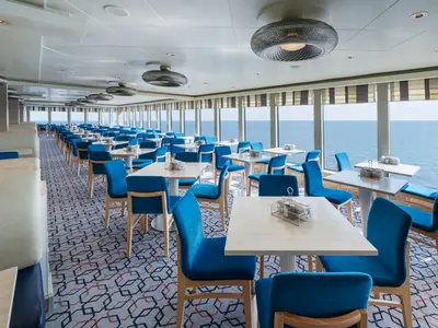Ein helles Restaurant an Bord der Norwegian Breakaway. Zahlreiche Tische und blaue Stühle sind zu sehen. Große Fenster bieten einen weiten Meerblick, während ein gemusterter Teppich den Boden ziert.
