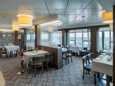 Elegantes Spezialitätenrestaurant Palomar an Bord der Norwegian Breakaway. Gedeckte Tische und Stühle sind zu sehen. Große Fenster bieten einen weiten Blick auf das Meer.