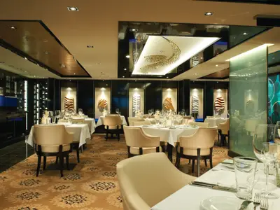 Elegantes Restaurant an Bord der Norwegian Breakaway mit stilvoll gedeckten Tischen, beigen Stühlen und modernem Dekor. Ein großer Kristallleuchter und Wandkunst schmücken den Raum. Ohne Personen.