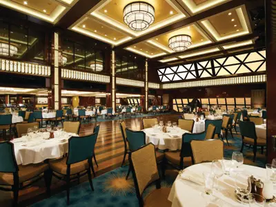 Blick in den grossen, eleganten Speisesaal "Manhattan Room" an Bord der Norwegian Breakaway, mit zahlreichen weiss gedeckten Tischen, teal-farbenen und goldenen Stühlen. Die hohen Wände sind mit dunklem Holz verkleidet und mit leuchtenden Paneelen und grossen Deckenleuchten ausgestattet. Im Hintergrund ist eine Bühne mit einem Klavier sichtbar.
