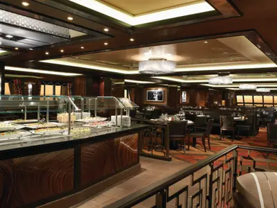 Buffet-Restaurant der Norwegian Breakaway mit langer Speisenauslage. Dahinter Tische und Meerblick durch Fenster. Elegantes Interieur, dunkles Holz, moderne Beleuchtung.
