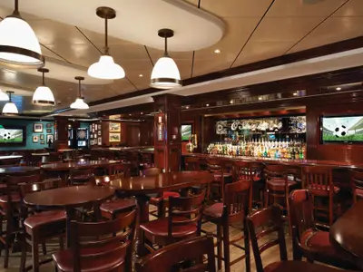 Innenansicht des O'Sheehan's Bar & Grill an Bord der Norwegian Breakaway. Gemütlicher Pub mit dunklen Holztischen, Stühlen, Barhockern und einem Tresen voller Spirituosen. Fernsehbildschirme zeigen Fussball.