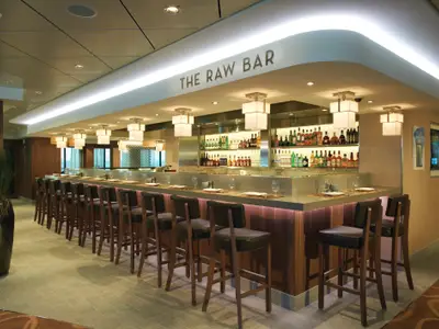 Blick in die Raw Bar an Bord der Norwegian Breakaway. Eine lange, moderne Bar mit Holzakzenten und vielen Barhockern. Im Hintergrund sind Spirituosen zu sehen.