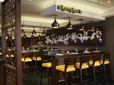 Das moderne Spezialitätenrestaurant Shanghai's an Bord der Norwegian Breakaway. Eine lange Bar mit Hockern und gedeckten Plätzen. Die Wand dahinter schmückt eine beleuchtete Kirschblütenkunst. Stilvolle Hängelampen und dunkle Holzelemente prägen das Interieur.