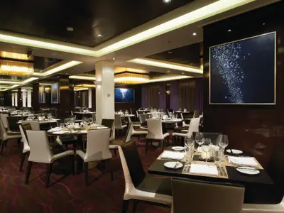 Elegantes Restaurant "Taste" an Bord der Norwegian Breakaway bei Abendstimmung. Zahlreiche gedeckte Tische, dunkles Holz, moderne Lichtleisten und Kronleuchter. Ein Kunstwerk mit Blasenmotiven an der Wand.