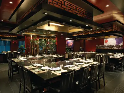 Das Teppanyaki-Restaurant an Bord der Norwegian Breakaway. Mehrere dunkle Tische mit Kochplatten und Stühlen sind bereit. Rote Wände, Bambuspflanzen und asiatische Dekorationen prägen das Interieur bei gedämpfter Abendstimmung.
