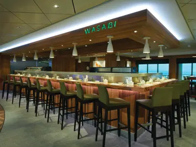 Innenansicht des Sushi-Restaurants Wasabi an Bord der Norwegian Breakaway. Eine lange Theke mit grünen Barhockern und modernen Hängelampen. Große Fenster bieten Meerblick.