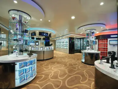 Heller, moderner Fotoshop an Bord der Norwegian Breakaway. Zylindrische Glasvitrinen präsentieren Kameras und Elektronik. Das Geschäft ist elegant beleuchtet und verfügt über einen gemusterten Teppich. Ohne Personen.