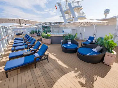 Blick auf das sonnige Sonnendeck der Norwegian Breakaway mit blauen Liegen, runden Daybeds, Sonnenschirmen und Whirlpool. Pflanzen und Holzdeck laden zum Entspannen ein.