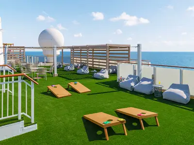 Ein sonniges Außendeck an Bord der Norwegian Breakaway mit Kunstrasen. Mehrere Cornhole-Spieltische und graue Sitzsäcke laden zum Verweilen ein. Im Hintergrund ist das blaue Meer zu sehen.