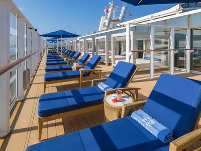 Sonniges Deck auf der Norwegian Breakaway mit Reihen blauer Lounge-Liegen und weissen Cabanas. Blaue NCL-Handtücher und bunte Cocktails auf Beistelltischen. Perfekt zum Entspannen.