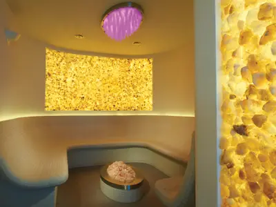 Detailansicht eines Spa-Salzraums an Bord der Norwegian Breakaway mit orange leuchtenden Salzkristallwänden und einer gemütlichen, geschwungenen Sitzbank. Eine Schale mit rosa Salzkristallen steht auf einem Tisch in der Mitte.