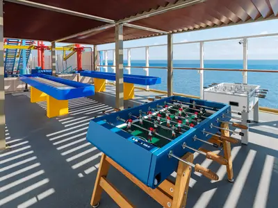 Ein offener Sportbereich an Bord der Norwegian Breakaway mit zwei Shuffleboard-Tischen und zwei Foosball-Tischen unter einer schattenspendenden Überdachung. Im Hintergrund ist das blaue Meer sichtbar.