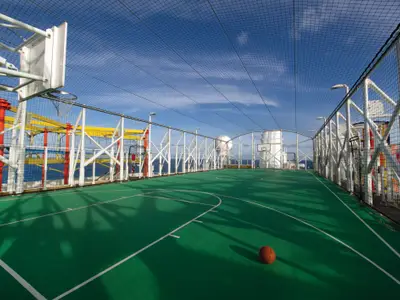Ein weitläufiger, grüner Basketballplatz unter blauem Himmel an Bord der Norwegian Breakaway. Ein Basketball liegt auf dem Vordergrund, umgeben von hohen Fangnetzen und einem farbenfrohen Hochseilgarten. Im Hintergrund ist das weite blaue Meer zu sehen.