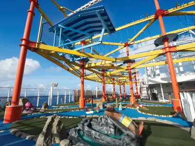 Der lebhafte Mini-Golf-Platz und der Hochseilgarten auf dem Oberdeck der Norwegian Breakaway. Orangefarbene Stützen, gelbe Balken und blaue Spielbahnen sind unter blauem Himmel mit Meerblick zu sehen. Das Deck ist menschenleer.