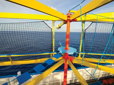 Blick auf das leuchtend gelbe und blaue Seilgarten-Element an Deck der Norwegian Breakaway, das über das offene, blaue Meer ragt. Ein Netz dient als Absicherung, der Himmel ist leicht bewölkt.