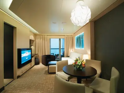 Geräumige Suite an Bord der Norwegian Breakaway mit hellem Wohnbereich, Fernseher, Esstisch, Sofa, Sesseln und einem Balkon mit Meerblick durch die offene Tür.