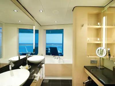 Geräumiges und helles Badezimmer einer luxuriösen Suite an Bord der Norwegian Breakaway. Es verfügt über zwei Waschbecken, eine Badewanne und ein großes Fenster mit Meerblick. Ein beleuchteter Vergrößerungsspiegel befindet sich auf dem Waschtisch.