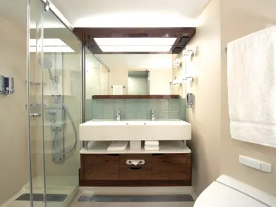 Modernes Badezimmer mit Glasdusche, Doppelwaschbecken und hellen Holzmoebeln an Bord der Norwegian Breakaway. Ein weisses Handtuch haengt rechts an der Wand.