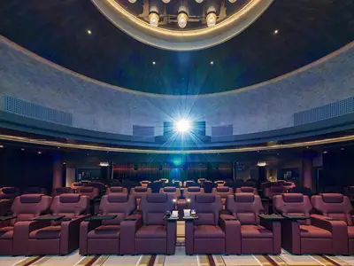 Blick in ein luxuriöses Kino an Bord der Norwegian Breakaway mit Reihen von roten Ledersitzen, Tischen mit Popcorn und einem hellen Filmprojektor.